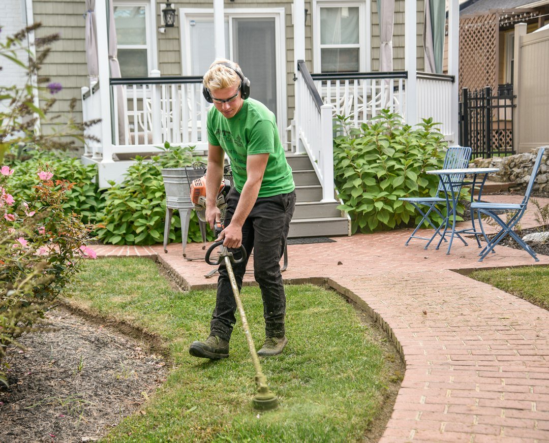 Lawn care worker trimming grass along a sidewalk in front of a customer's house, comparing Jobber vs Housecall Pro for lawn care businesses